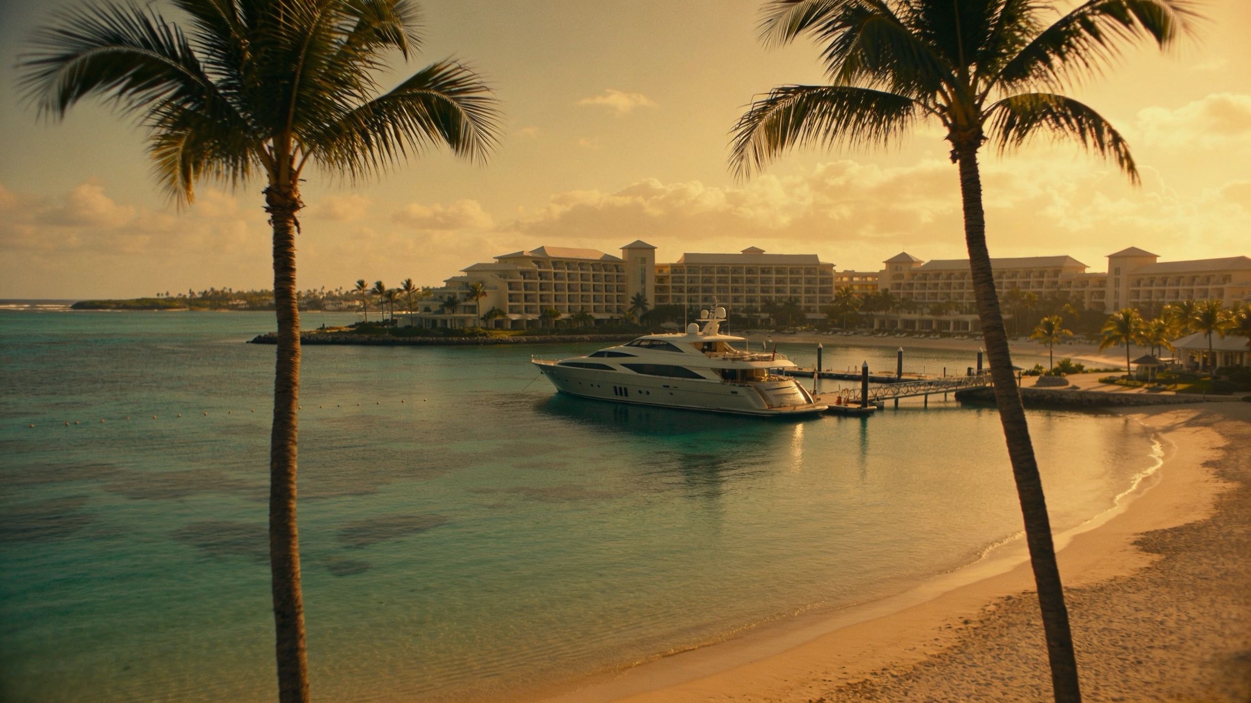 A luxurious white yacht docked in a tropical bay near a large resort hotel at sunset.